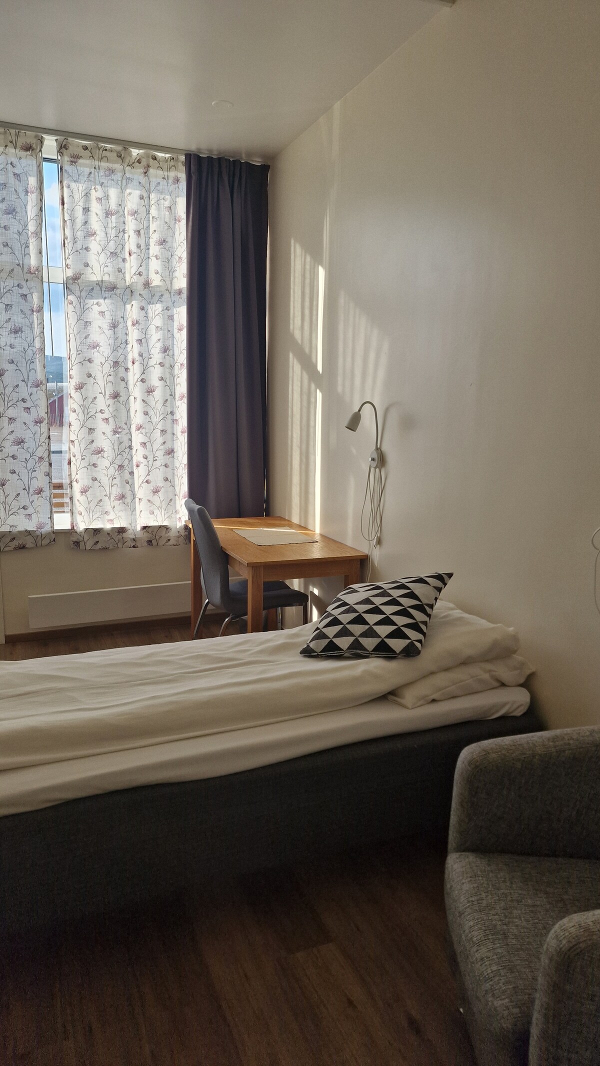 A comfortable sleeping area contains a single bed with white bedding. A chair and desk are positioned by a large window, adorned with floral-patterned curtains. Natural light fills the space, highlighting the warm tones of the wooden floor and the gray accent chair.