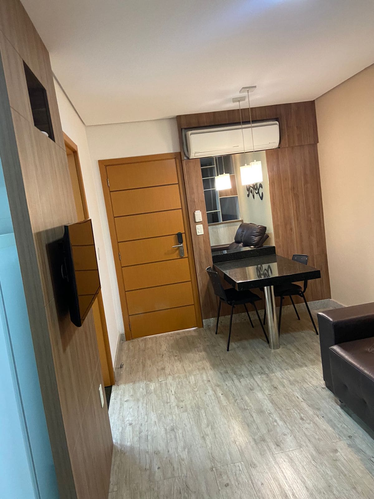 A dining area features a glass table with four black chairs, complemented by a modern light fixture. The room showcases warm wooden accents, a brown sofa, and an air conditioning unit mounted on the wall. A door provides access to the hallway.