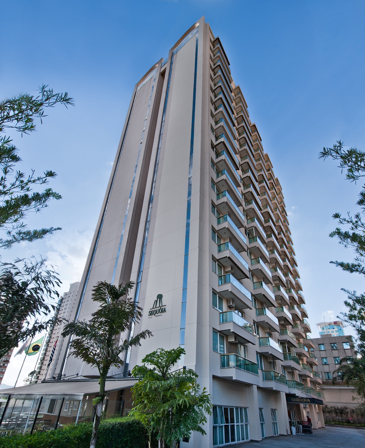 A tall modern building is set against a clear blue sky, featuring multiple floors with balconies. Lush greenery is visible at the base, framing the entrance, which is marked by the hotel's signage. The design emphasizes a sleek and contemporary architectural style.