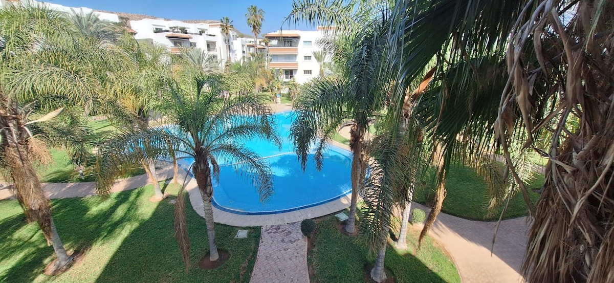The image showcases a serene swimming pool surrounded by lush palm trees and manicured grass. A pathway winds around the pool, leading through the landscaped garden. Sunlight reflects off the water's surface, creating a calming atmosphere.
