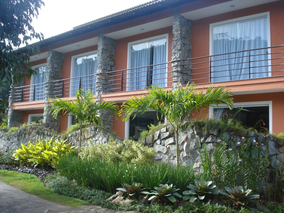 The image showcases a serene building featuring multiple large windows framed by white curtains and a balcony with a railing. Lush greenery, including ornamental plants and flowering shrubs, surrounds the stone-faced structure, creating a natural and inviting atmosphere.