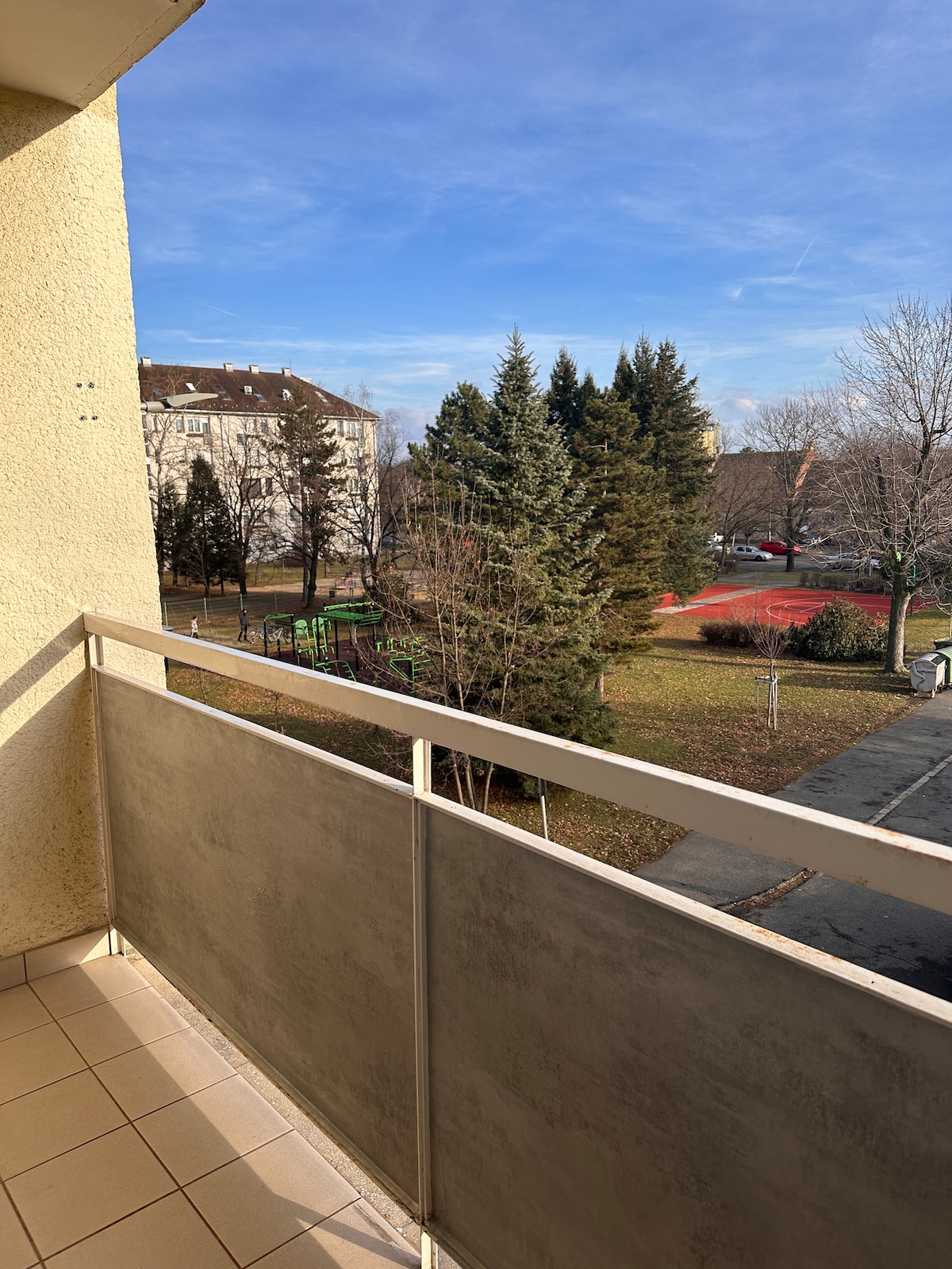 A private balcony overlooks a green park area featuring tall trees and a playground. Nearby, a sports court is visible, providing recreational space. The sky is bright, and the surrounding buildings add a sense of community to the view.