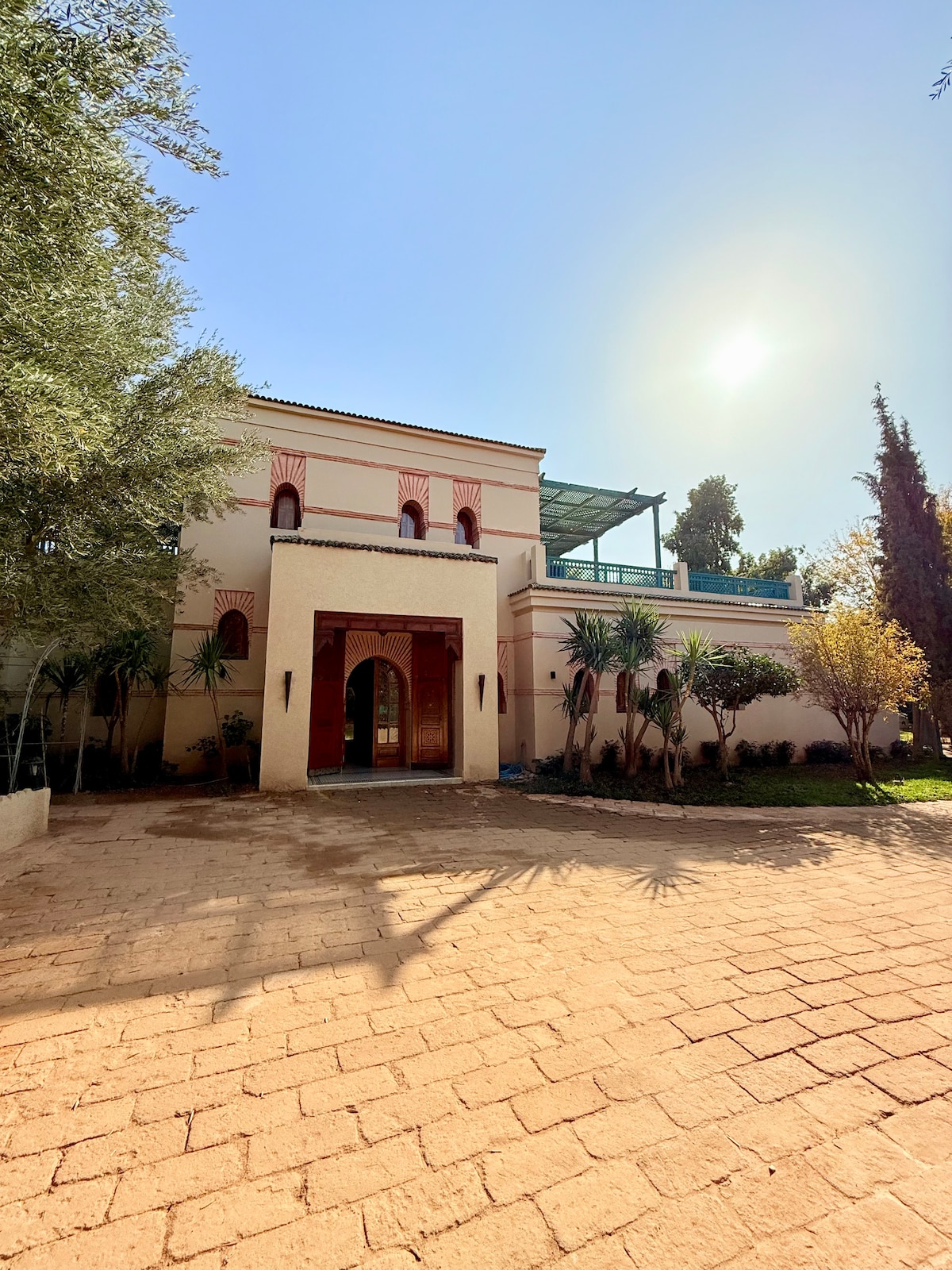 The villa's exterior features a welcoming façade with arched doorways and a landscaped garden. Sunlight illuminates the structure, casting shadows across the courtyard paved with terracotta tiles. Lush greenery and palms surround the property, enhancing its serene setting.