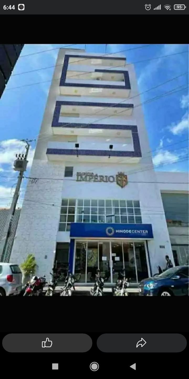 The exterior view of a modern building showcases a clean, white facade with blue accents. The entrance features large glass doors labeled 'HINODE CENTER,' flanked by several parked motorcycles and vehicles. The building has multiple floors, clearly visible with a horizontal arrangement of windows.