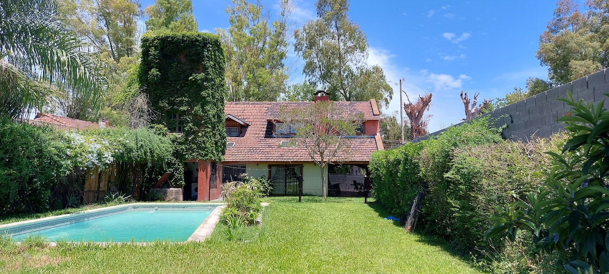 A spacious garden surrounds a clear swimming pool, with lush greenery and trees providing shade. The two-story house with a charming roof is partially obscured by climbing vines, while a bright blue sky is visible above.