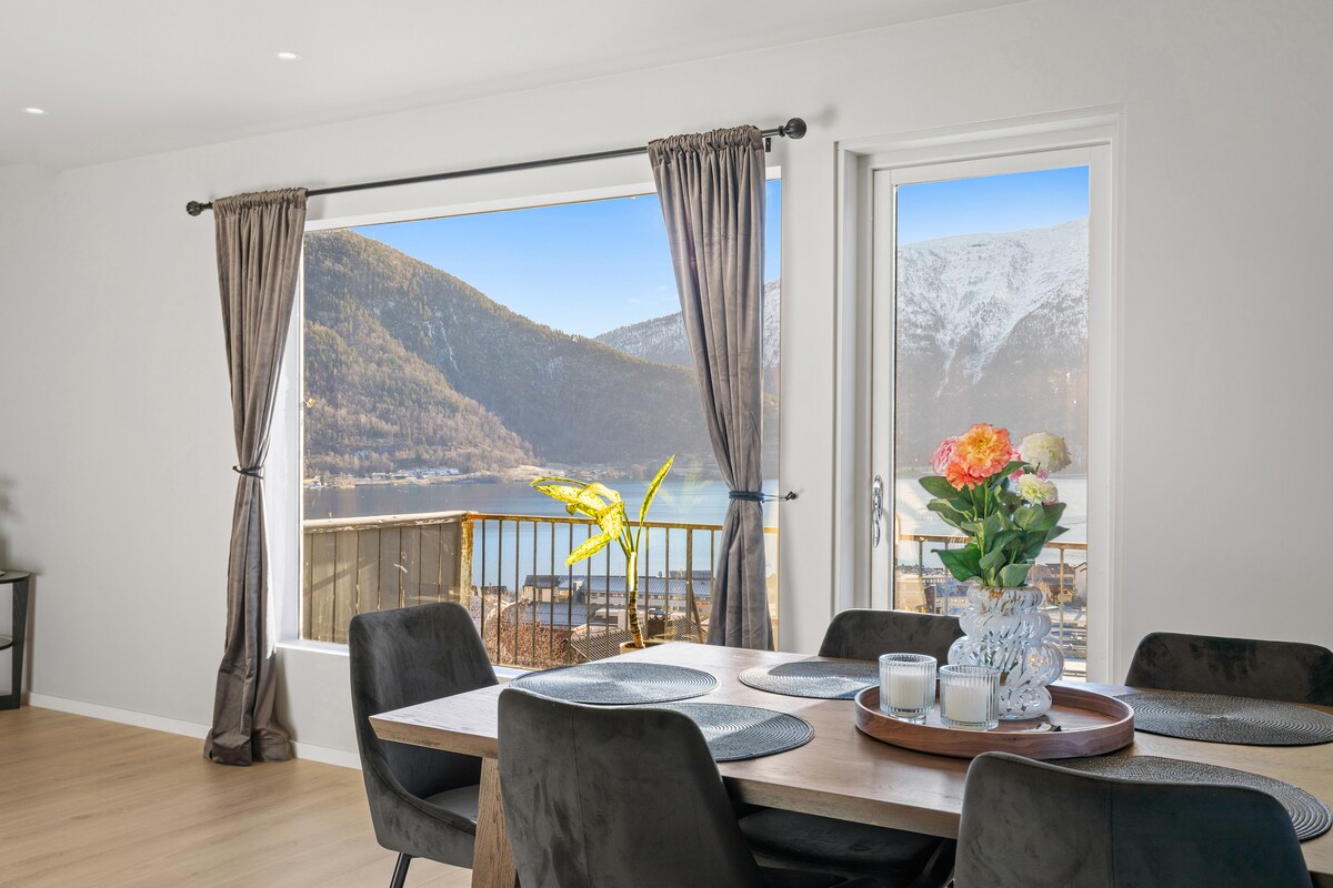 A dining area is presented with a wooden table surrounded by dark chairs. Large windows offer a stunning view of the mountains and water outside, while natural light fills the space. A vase with fresh flowers sits on the table, adding a touch of color.