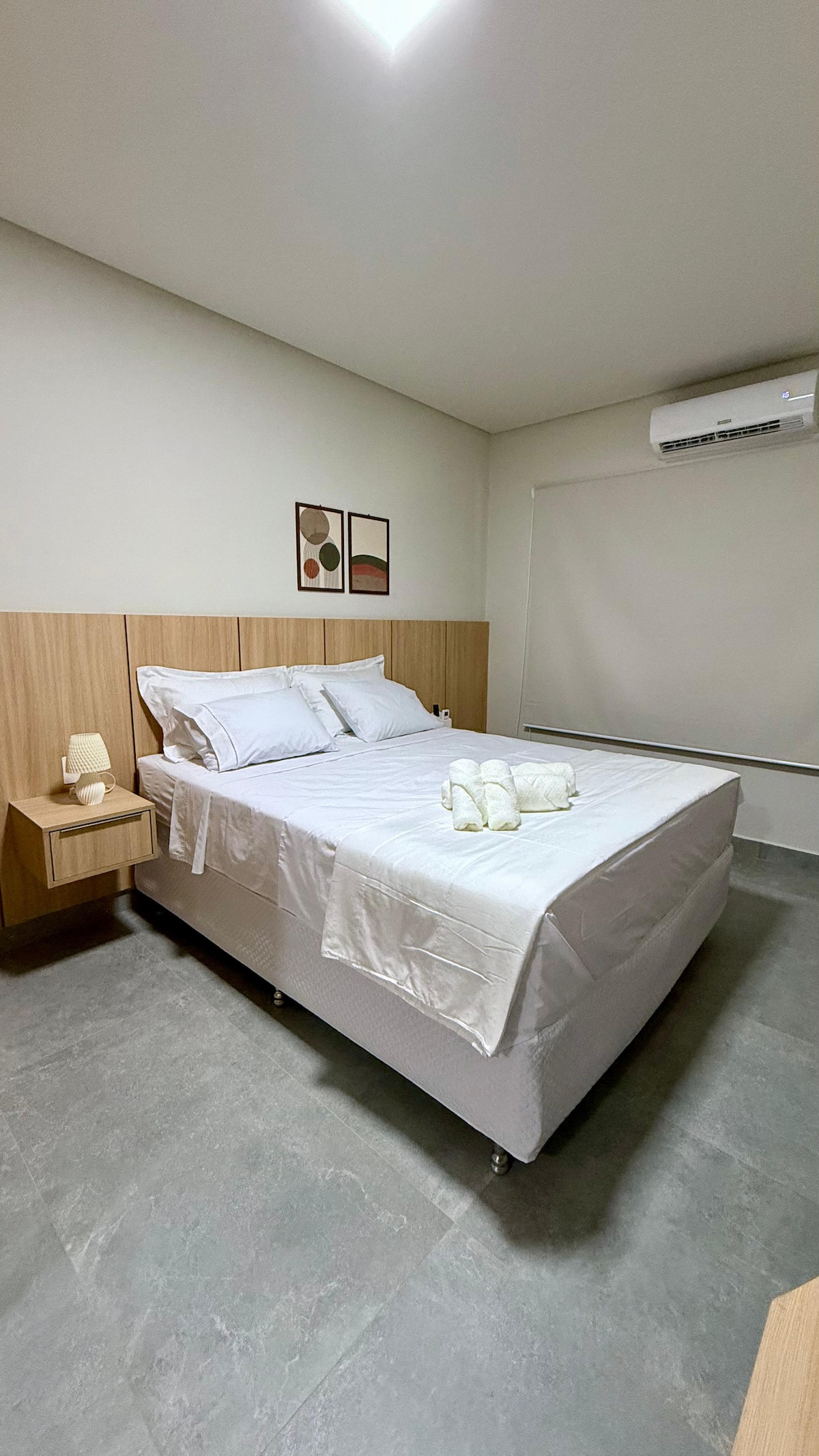 A cozy bedroom features a spacious bed with fresh white linens and towels neatly arranged on top. Soft lighting is provided by a bedside lamp on a wooden nightstand. The decor includes minimalistic artwork, while a window covered by a blind allows for natural light.