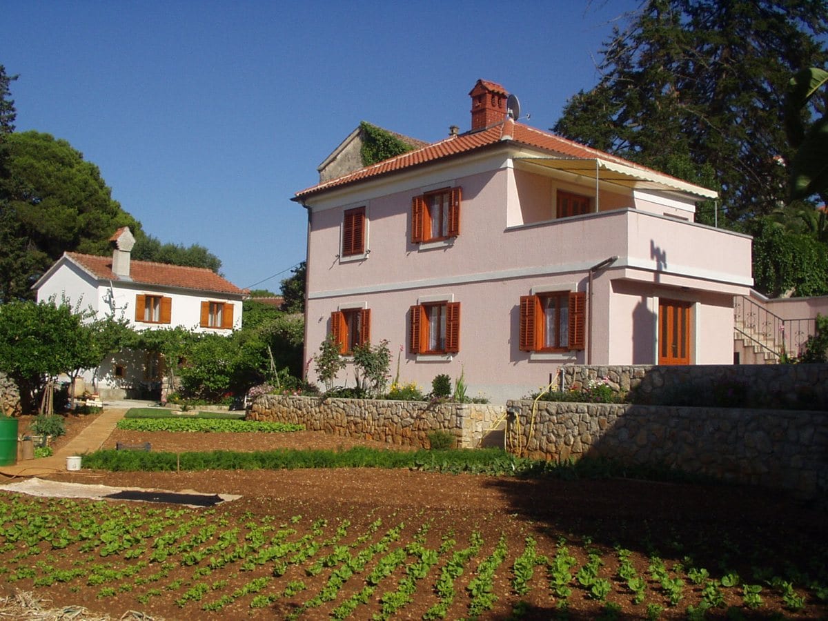 A two-story house is set within a spacious garden, featuring a well-maintained vegetable patch in the foreground. The exterior is painted a soft pink, with orange window shutters adding a vibrant touch. Lush greenery surrounds the property, enhancing the tranquil atmosphere.