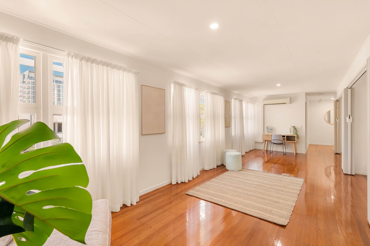 A light-filled living area features large windows with sheer white curtains, offering a bright ambiance. Wooden floors provide a warm touch, while a comfortable sofa and a light-colored rug create a cozy seating space. A small dining table with chairs is positioned nearby, enhancing functionality.