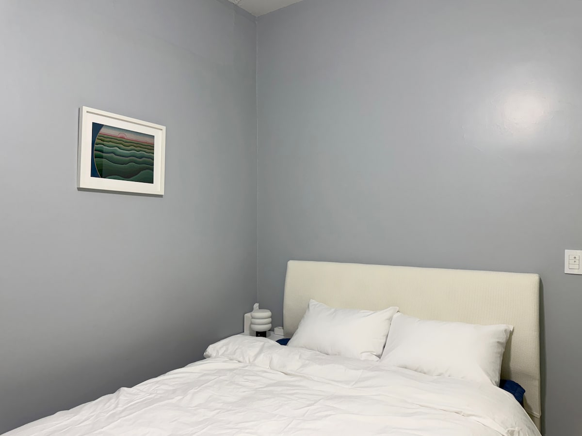 A cozy bedroom is depicted, featuring a queen-sized bed with white linens and multiple pillows. A minimalist lamp is positioned on the bedside table, and a piece of wall art adds a splash of color to the muted gray walls.