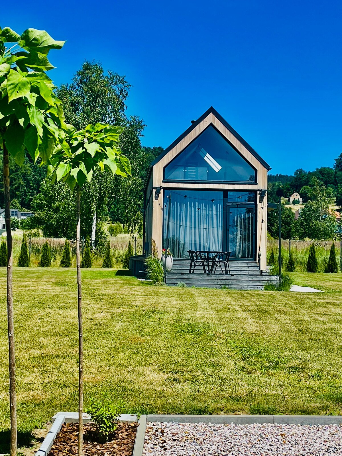 A modern cottage features a sloped roof and large glass front, bringing natural light indoors. A small deck with a seating area is visible, embraced by a well-maintained lawn and young trees, surrounded by scenic greenery.