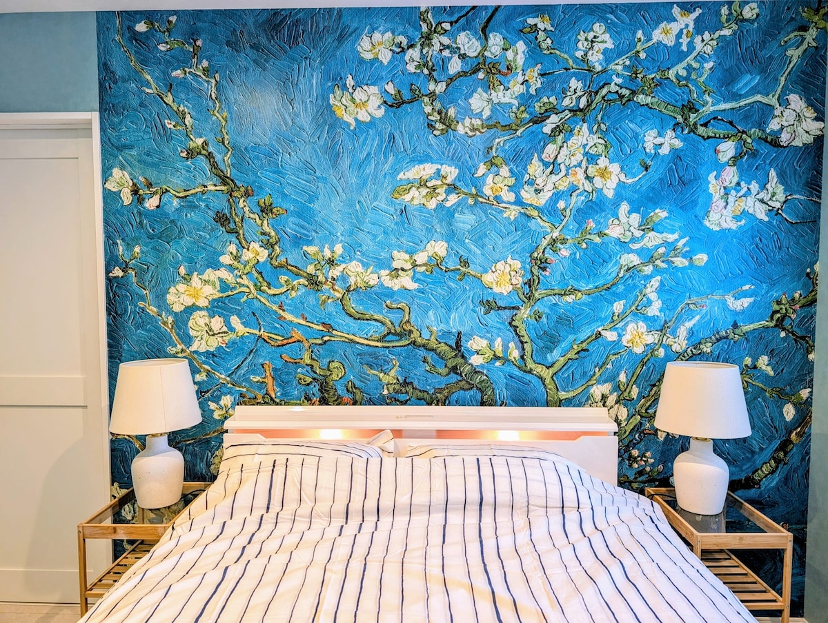 A serene bedroom features a striking blue wall adorned with a floral mural. Soft bedding is arranged neatly on the spacious bed. Two matching bedside lamps with warm light are placed on either side, complementing the tranquil ambiance created by the overall decor.