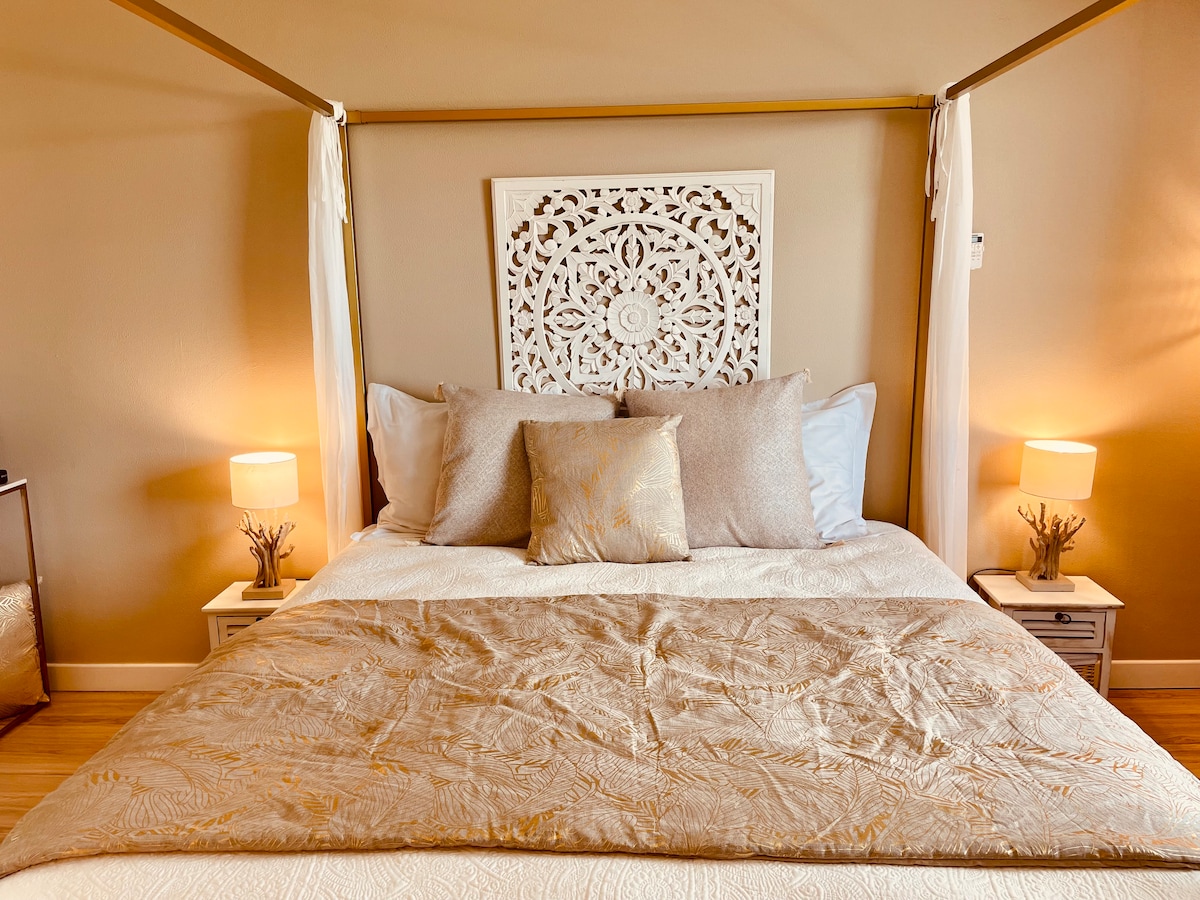 A king-size canopy bed is centered in the room, adorned with neutral-toned bedding and decorative pillows. Two bedside lamps provide soft illumination, while an intricately carved headboard adds a touch of elegance to the space.