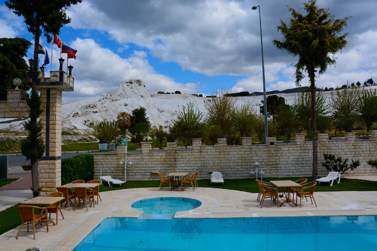 A swimming pool is surrounded by well-maintained green grass and lounge chairs. In the background, white travertine terraces rise against a cloudy sky, with trees and shrubs lining the edges. Flags are displayed near the pool area, adding a touch of character to the setting.