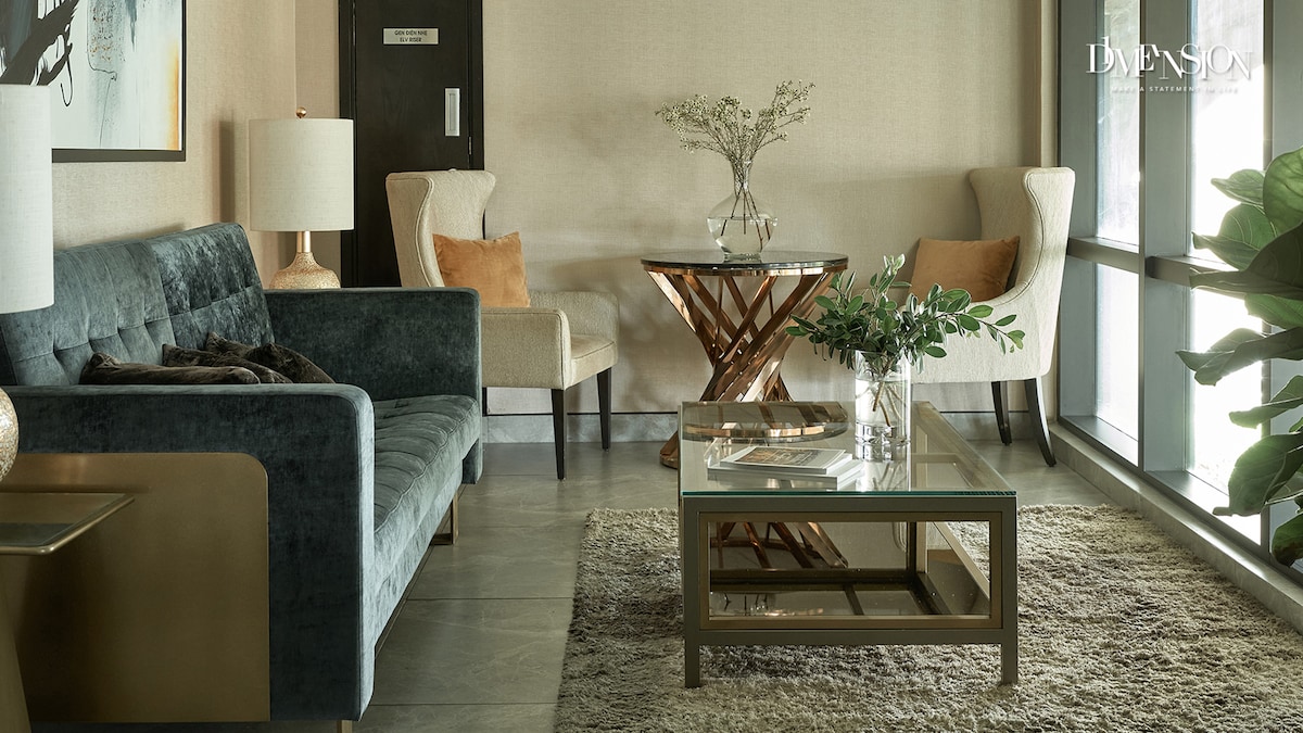 The living room features a stylish seating area with a plush blue sofa and two cream-colored chairs. A wooden side table with a geometric base stands beside a glass coffee table, both adorned with decorative plants. Natural light filters through large windows, enhancing the warm, neutral tones.