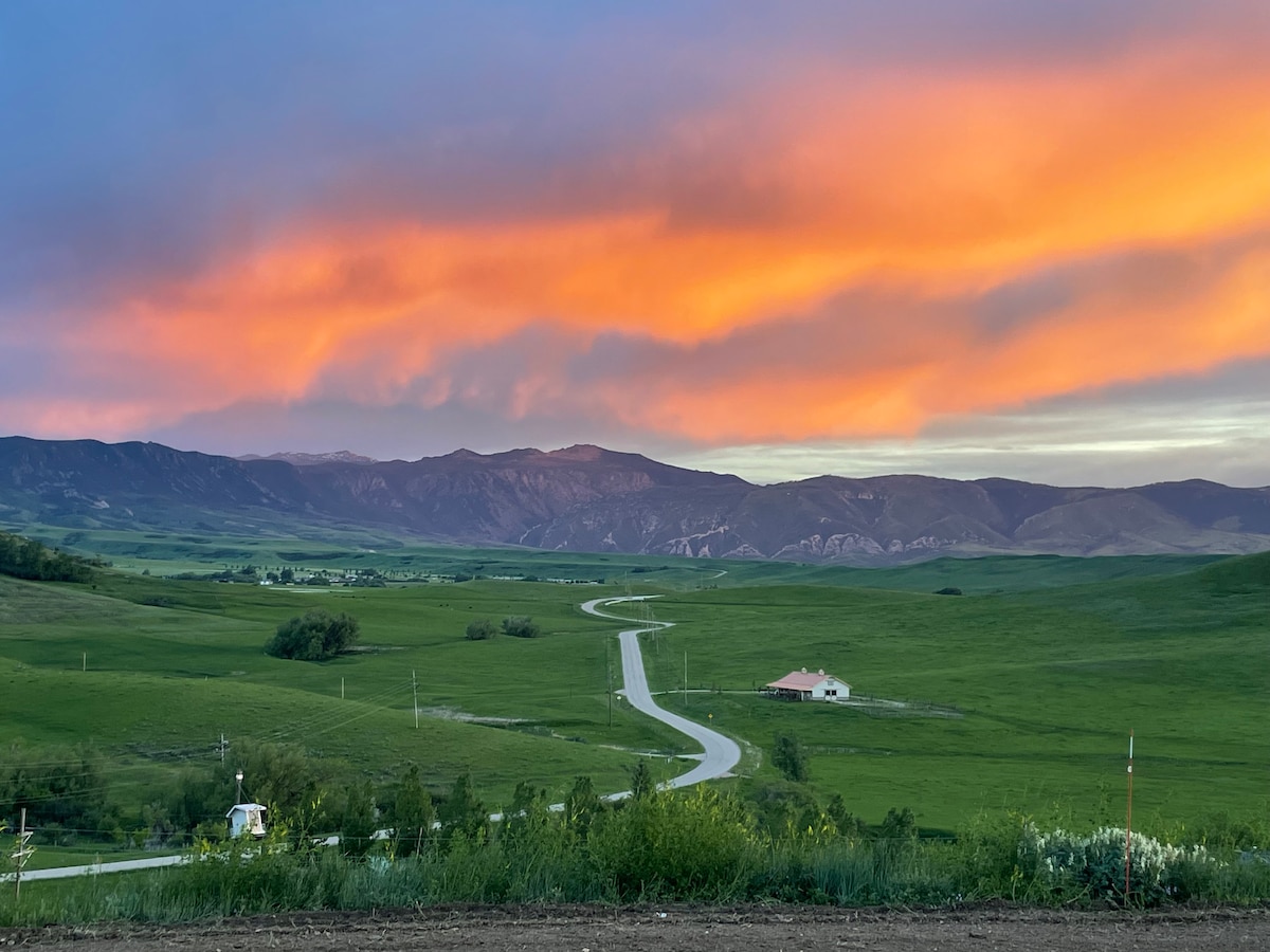 A panoramic view of rolling green hills stretches toward the horizon, with a winding road leading through the landscape. The sky is painted in shades of orange and pink during sunset, highlighting the silhouette of distant mountains.
