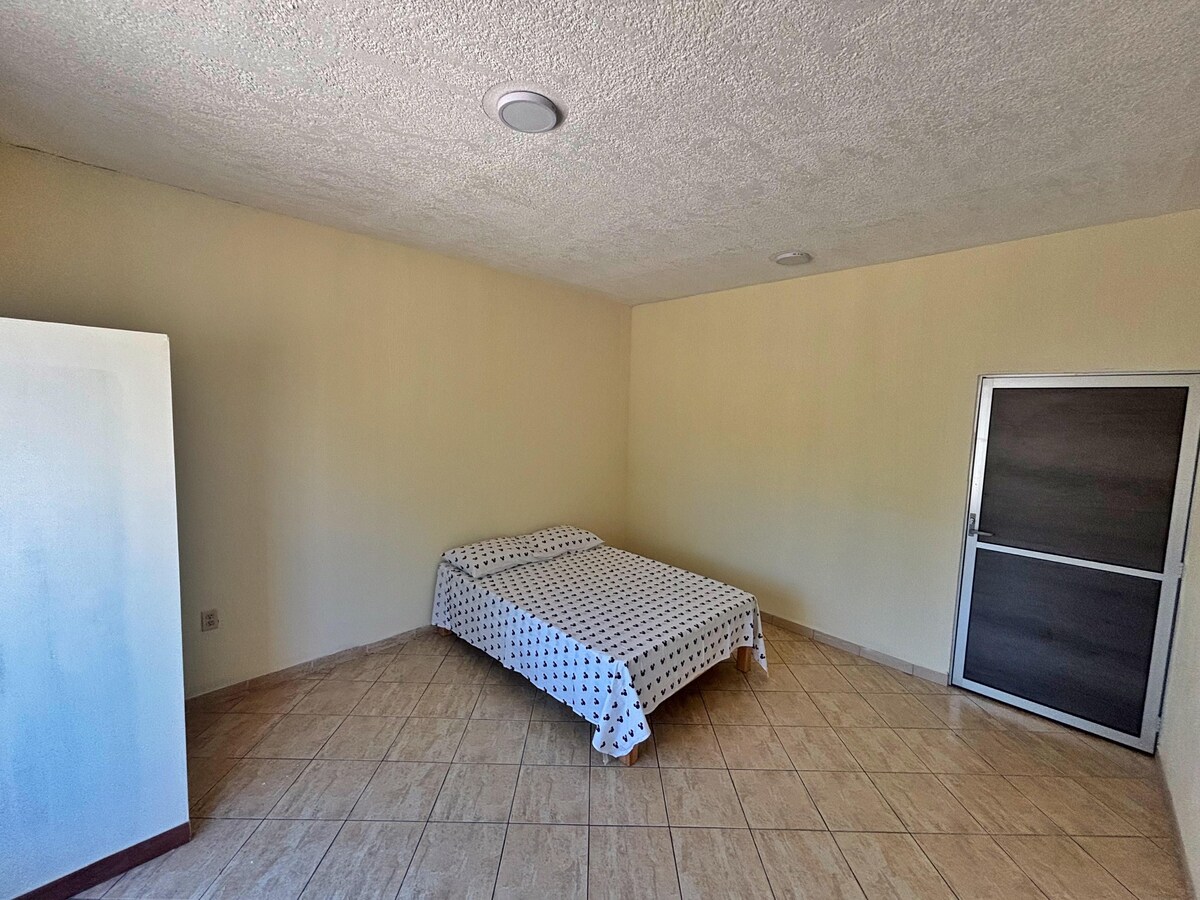 A spacious room features neutral-colored walls and tiled flooring. A bed with a patterned cover is positioned against one wall. A simple wardrobe stands opposite the bed, while a glass door is visible, leading to another area.