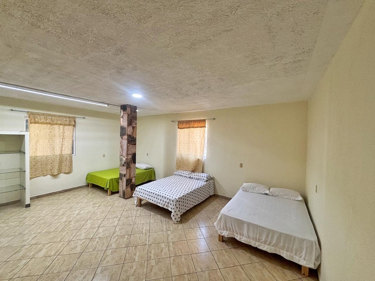 A spacious room is furnished with three beds, including a green bed with a solid cover, a patterned bedspread, and a bed with white linens. Natural light enters through two windows adorned with light curtains. The floor features neutral tiles, enhancing the openness of the space.