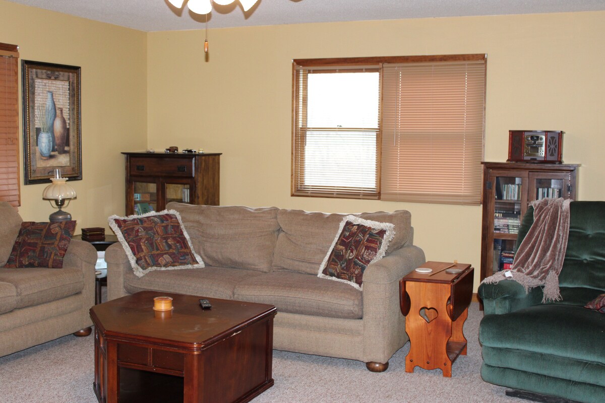A comfortable living area features a spacious sofa with decorative pillows and a green armchair. A wooden coffee table with a heart-shaped design sits in the center, surrounded by a light-colored carpet. Natural light enters through two windows adorned with blinds.