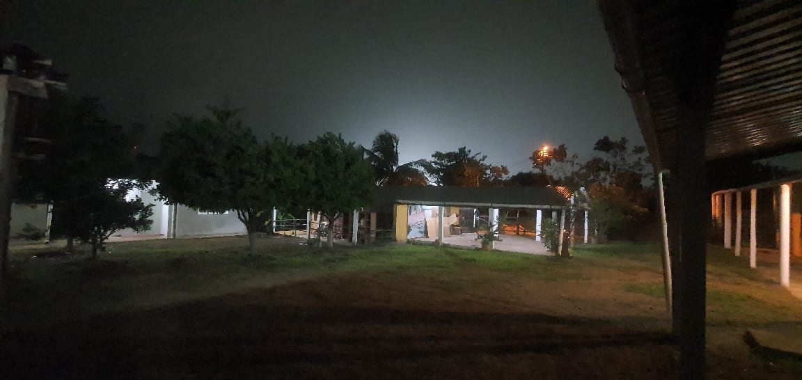 A dimly lit outdoor area is surrounded by lush greenery, offering a tranquil setting. A covered space is visible, with several wooden benches and tables arranged beneath it. Soft light from the nearby property casts a gentle glow, creating a serene evening atmosphere.