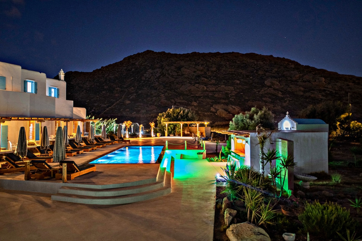 An outdoor pool area is illuminated in green and blue lights, surrounded by lounge chairs and lush gardens. The backdrop features a prominent mountain and an adjacent structure, enhancing the serene ambiance. Various plants and trees are visible near the poolside.