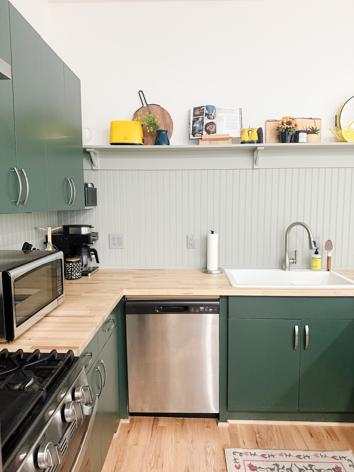 A compact kitchen design features green cabinetry and wood countertops. Stainless steel appliances, including a dishwasher and oven, are visible. A well-organized shelf holds decorative items and plants. Natural light illuminates the space, highlighting the soft green paneling above the sink.