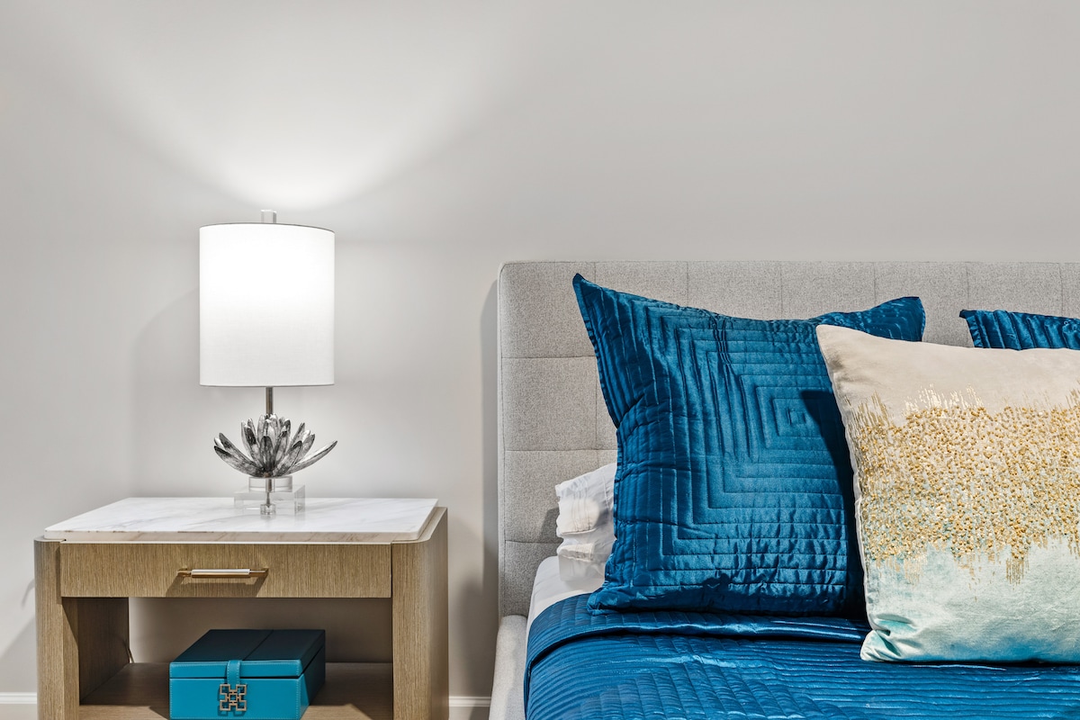 A well-appointed bedroom features a plush bed adorned with textured blue and gold pillows. A modern lamp casts a soft glow from a stylish bedside table, which includes a decorative blue box. The light color palette contributes to a serene atmosphere.