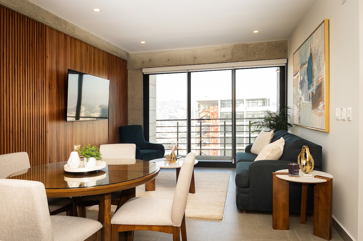 A stylish living area is highlighted by a wooden accent wall and large windows offering city views. Comfortable seating includes a dark blue sofa and a circular dining table surrounded by light fabric chairs. A mounted television is visible, enhancing modern amenities.