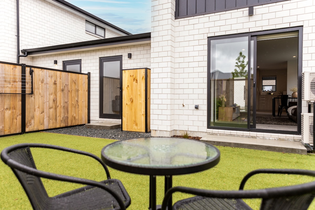 A private outdoor space is displayed featuring a small glass table surrounded by two black chairs. The area is bordered by wooden fences, with a well-maintained green lawn visible. Open glass doors provide a view into the indoor living area.