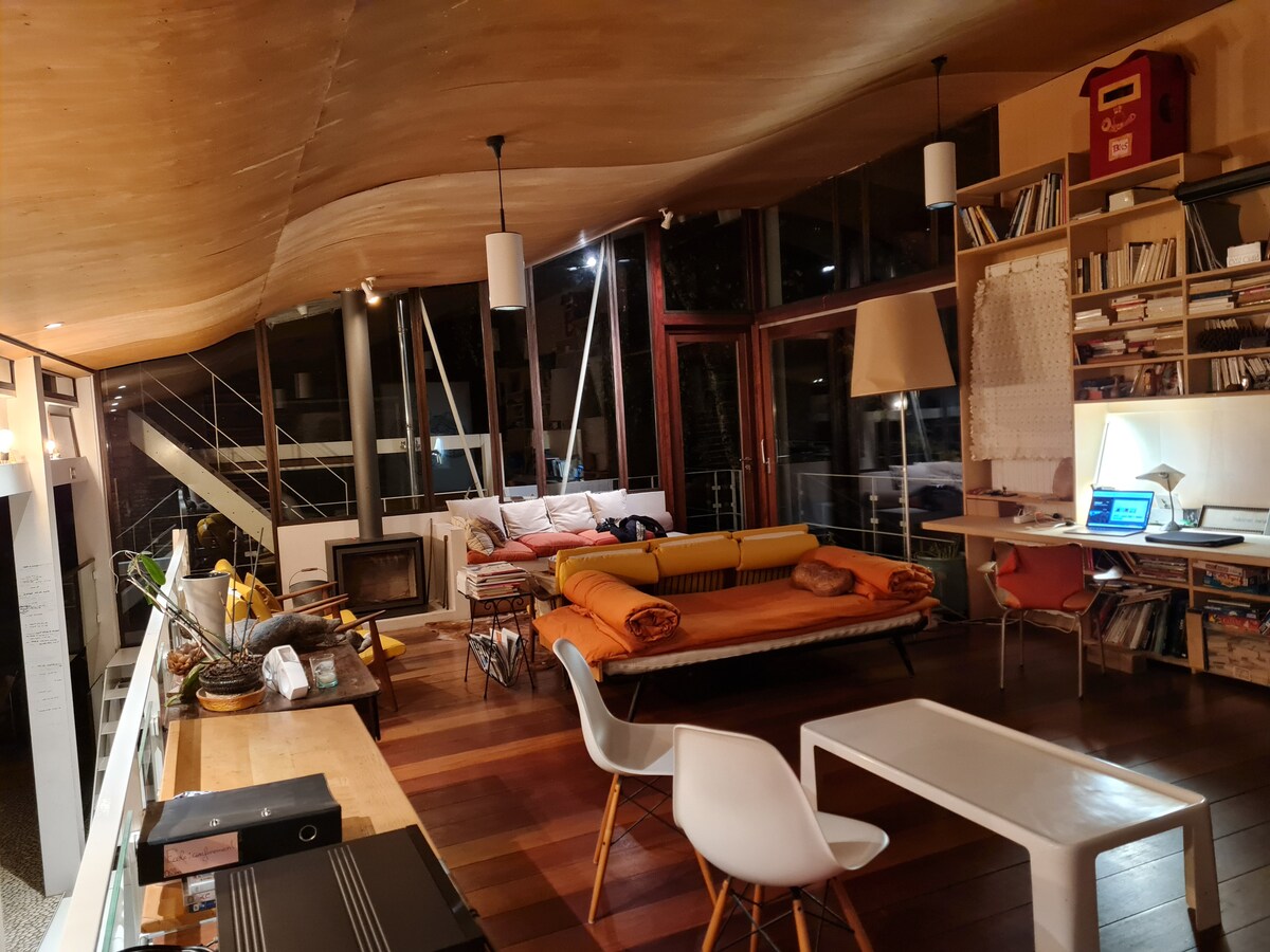 The living area features a unique curved wooden ceiling and large windows that allow natural light to fill the space. A mix of seating options, including a sofa and dining table, is present. A modern workspace with a computer sits against the wall.