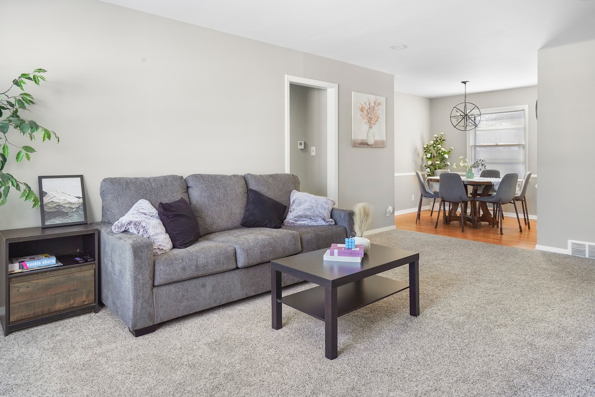 A spacious living area features a large gray sofa accompanied by a low coffee table. Simple decor includes a plant and a framed picture on the side table. A doorway leads to a dining area with a modern light fixture and a round table set for six.