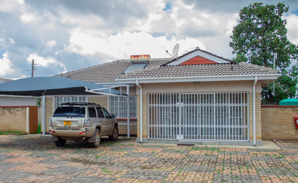 The exterior of the home features a well-maintained façade with a grey roof and white walls. A secure gated entrance is evident, along with a spacious driveway accommodating a parked vehicle. The surrounding area includes subtle landscaping and a shaded carport.