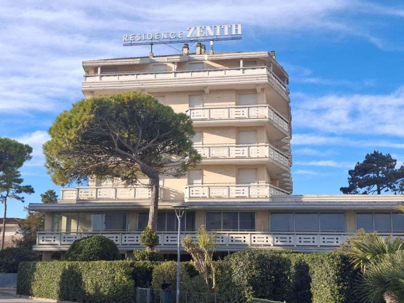 The building features a modern architectural design with multiple stories and expansive windows. The name 'Residence Zenith' is prominently displayed at the top. Surrounding greenery includes tall trees and shrubs, providing a serene environment. A clear blue sky enhances the view.