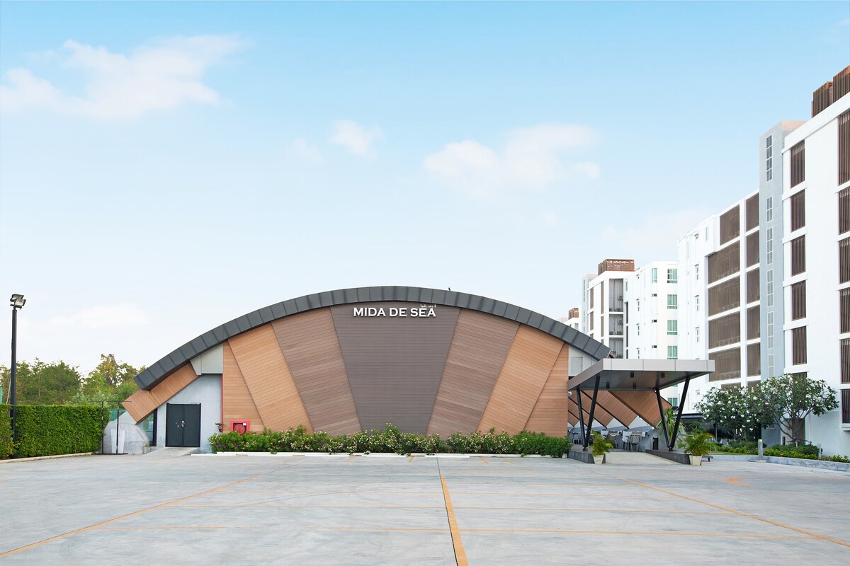 The exterior of Mida De Sea resort is displayed, showcasing a modern building with a distinctive curved design. A clear blue sky is visible overhead, and the surrounding area is well-maintained, featuring landscaped greenery and a spacious parking lot.