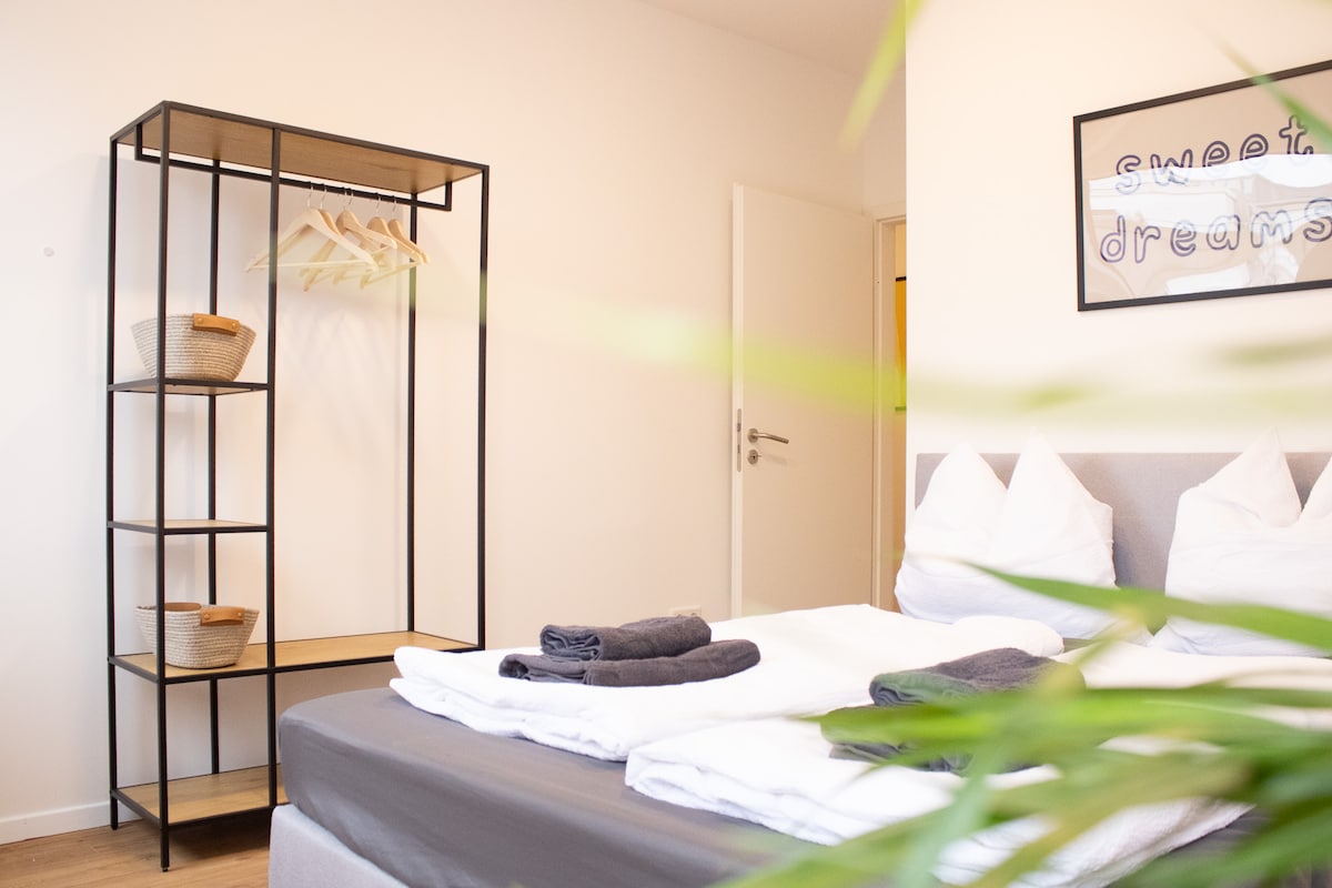 The sleeping area features a spacious bed with neatly arranged blankets and towels. A minimalist clothing rack stands nearby, displaying baskets. A potted plant adds a touch of greenery, while a wall decoration conveys a positive message.