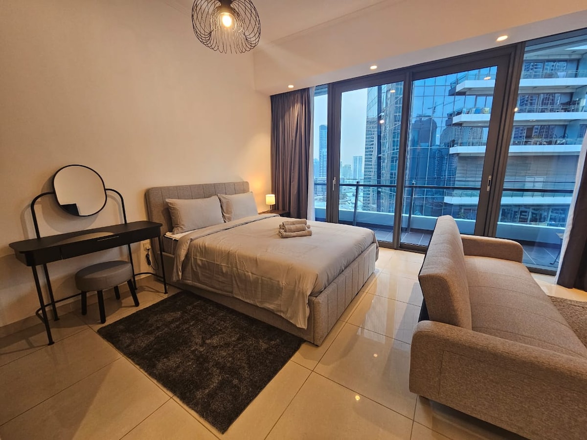 A modern studio features light-colored furnishings and floor-to-ceiling windows offering views of the Dubai skyline. The inviting sleeping area contains a cozy bed with soft linens beside a minimalist desk and chair. A neutral-toned area rug complements the tiled floor, while a comfortable sofa completes the space.