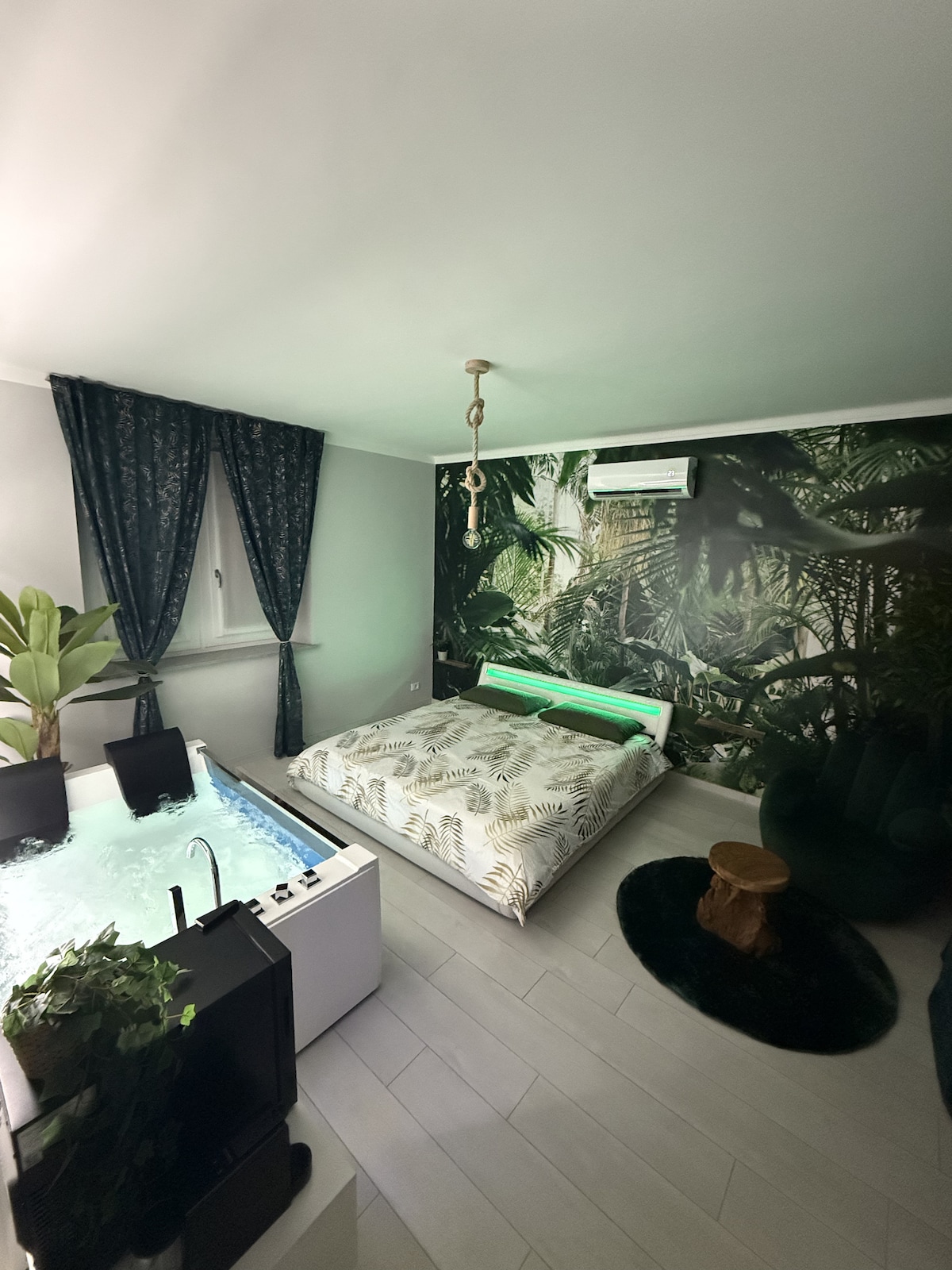 The room features a spacious bed adorned with tropical-patterned bedding, complemented by a large green area rug. A transparent Jacuzzi bath is positioned nearby, with potted plants adding a touch of nature. Dark curtains frame a window, providing a serene ambiance.