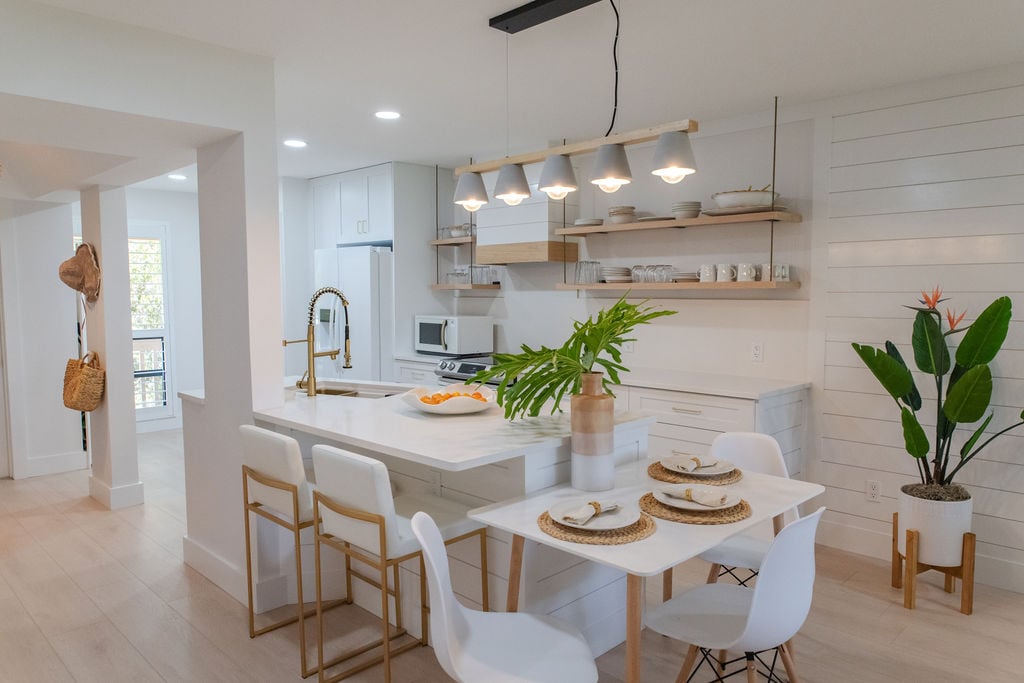 A modern kitchen and dining area is presented, featuring a sleek white table set for four with gold accents. A stylish pendant light hangs above, while a potted plant adds a touch of greenery. White cabinetry and counter space are complemented by new appliances in the background.