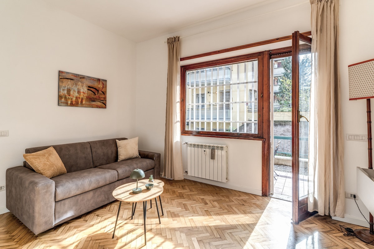 A spacious living area features a modern sofa with cushions, complemented by a circular coffee table. Natural light fills the room through large windows, showcasing herringbone wood flooring. Access to a private balcony is provided by glass doors, enhancing the airy atmosphere.