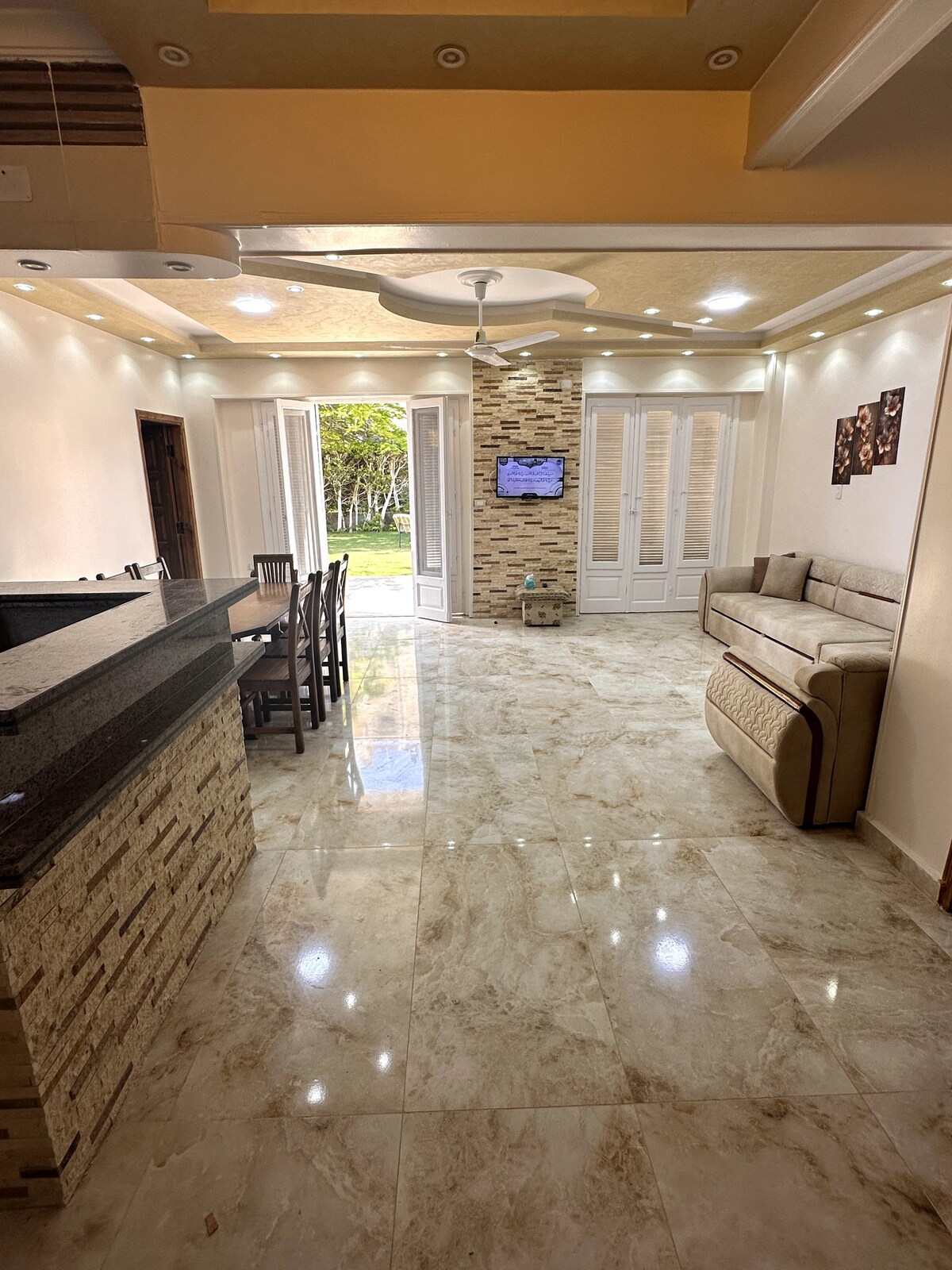 An open living area is presented, featuring polished marble flooring that reflects light. A comfortable sofa is positioned next to a dining area with multiple chairs, while large glass doors lead to a view of outdoor greenery. Soft lighting enhances the space's inviting ambiance.
