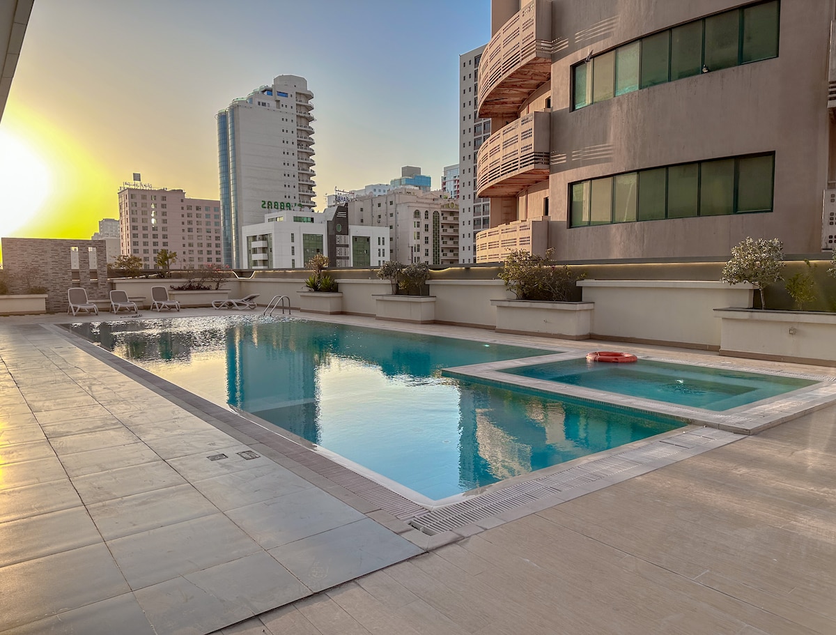 A serene swimming pool area is bathed in golden light from the setting sun, reflecting on the water's surface. Surrounding the pool, modern architecture displays a mix of residential buildings. Comfortable lounge chairs line the poolside, inviting relaxation in this tranquil outdoor space.
