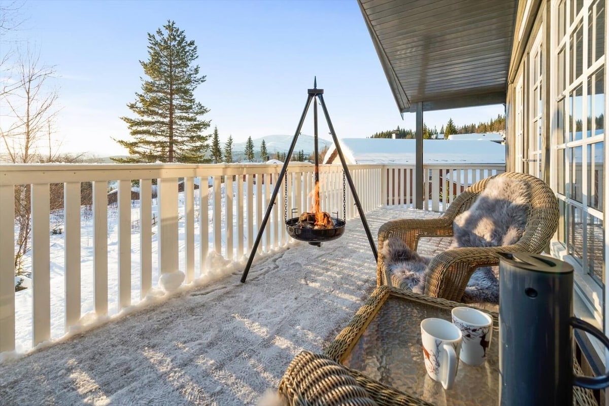 A spacious balcony features a cozy seating area with woven chairs and a table, set against a winter landscape. A modern fire pit is centered on the balcony, providing warmth and ambiance. Snow-covered ground and distant mountains are visible, illuminated by natural light.