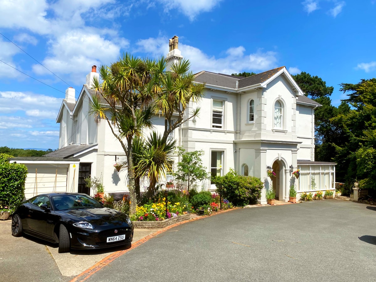 A grand Victorian villa features a fresh white façade framed by lush greenery and vibrant flower beds. In the foreground, a sleek black car is parked on a winding driveway that leads to the entrance, under a clear blue sky dotted with white clouds.
