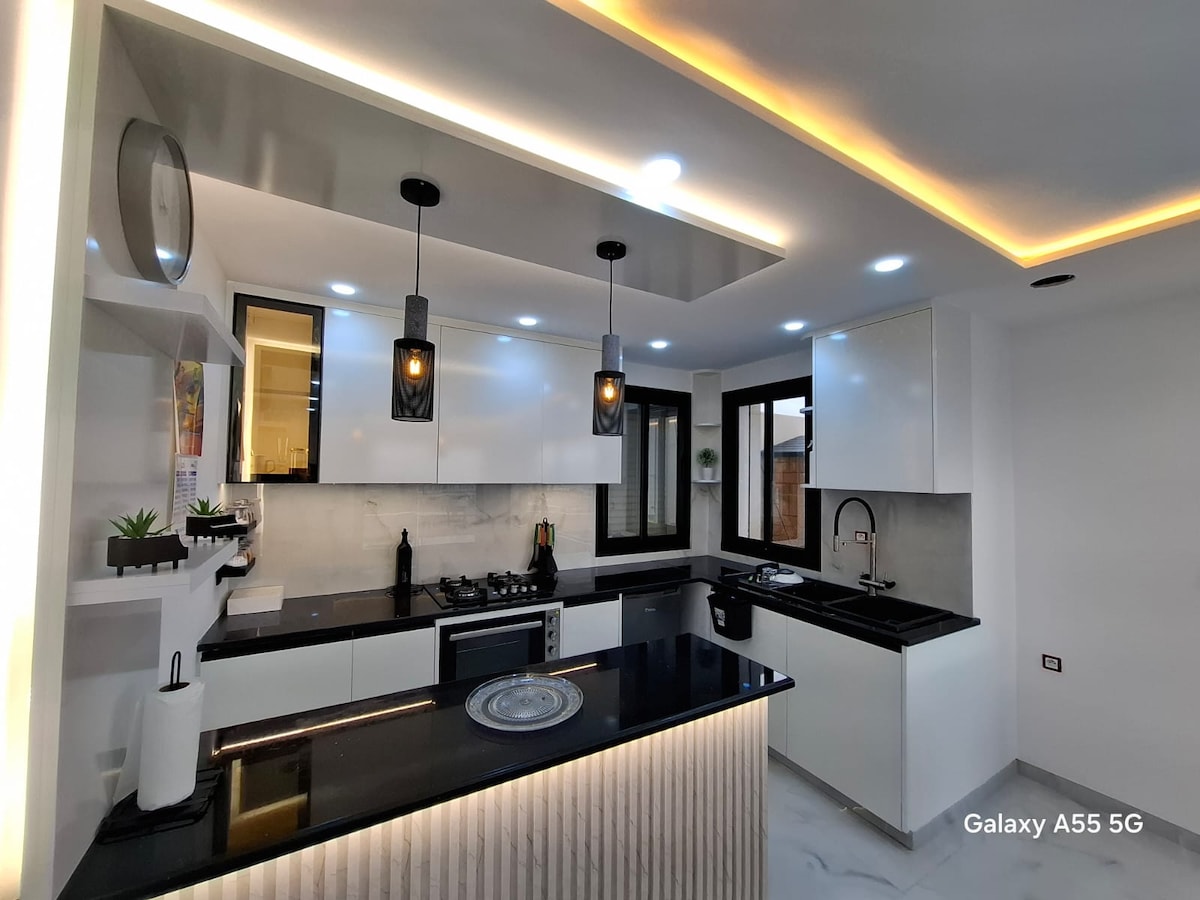 A modern kitchen is featured with sleek black countertops and white cabinetry. Task lighting is provided by stylish pendant lights hanging above the central island. A window allows natural light, and essential appliances are easily accessible, creating a functional cooking space.