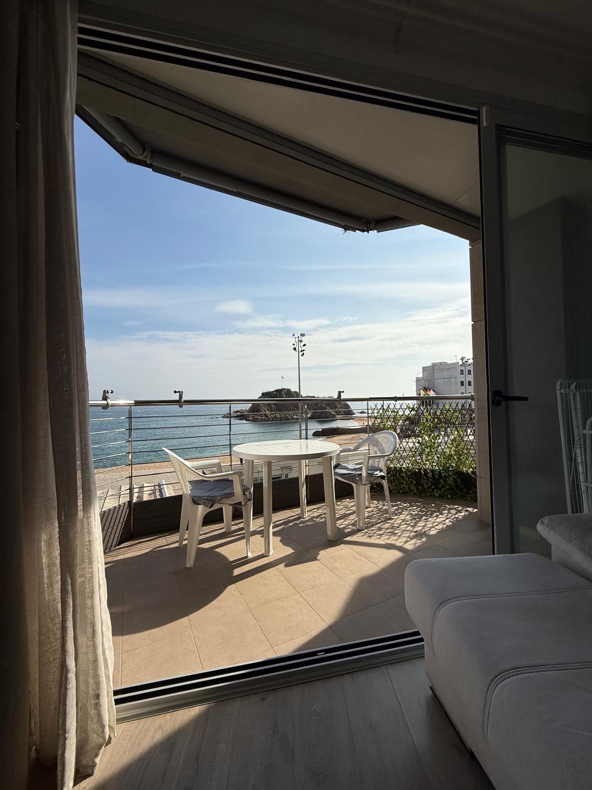 A terrace is shown overlooking the sea, featuring a table and chairs for outdoor dining. The water reflects the soft light of the sky, framed by a simple railing. Nearby, coastal architecture is visible, contributing to the serene view.