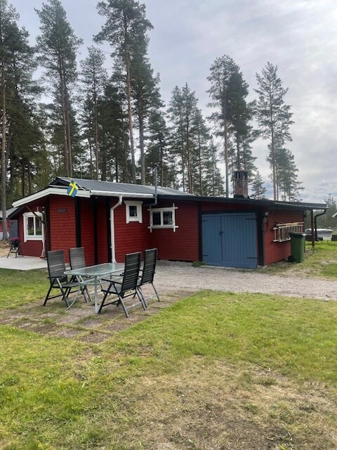 A red house is surrounded by tall trees and a cleared lawn. An outdoor dining area with a table and six chairs is positioned on a gravel path, while a detached garage is visible beside the house. A Swedish flag is displayed near the house.