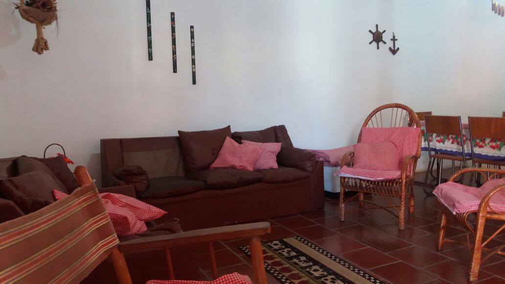 A casual living space features a dark brown sofa adorned with light pink cushions. Several woven chairs, also with pink coverings, are arranged around a wooden table. Decorative wall elements, including fish and anchors, provide a coastal touch to the room.