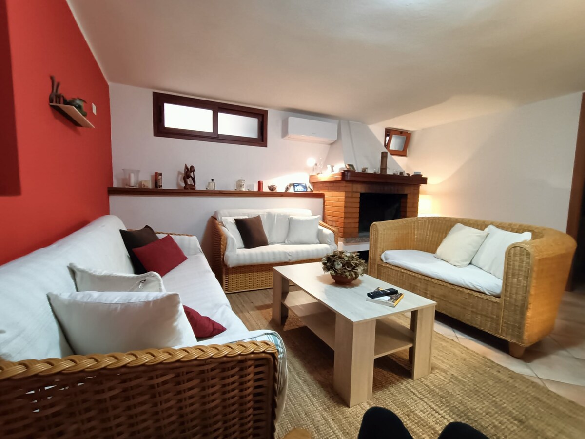 A cozy living space features comfortable rattan seating arranged around a light wooden coffee table. A warm fireplace is positioned on the wall, and natural light enters through windows, enhancing the relaxing atmosphere of the room.