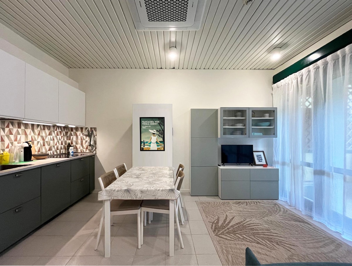 The bright living area features a modern, fully equipped kitchen with gray cabinetry and a decorative backsplash. A dining table for four sits center stage under soft lighting, surrounded by stylish chairs. A media unit with a flat-screen TV is positioned nearby, enhancing the organized layout.