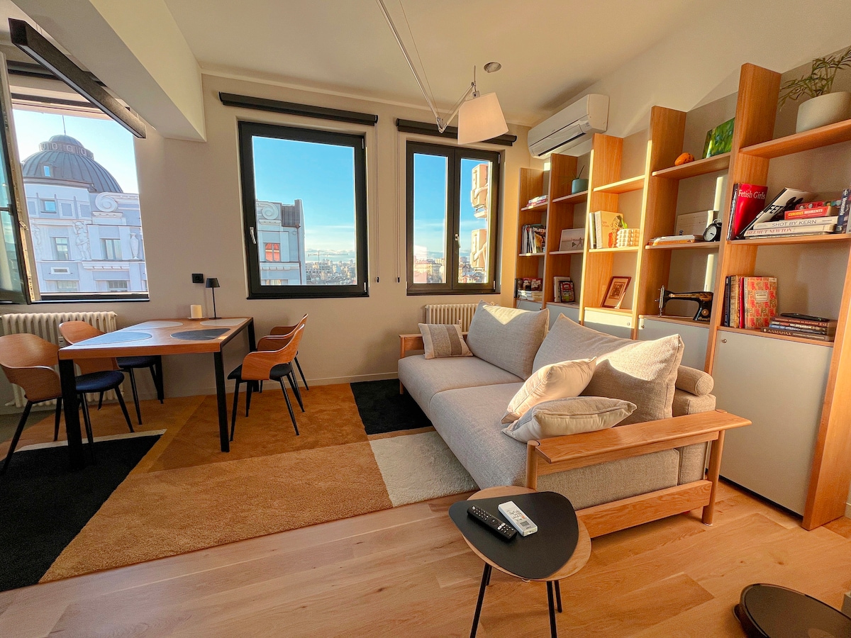 A cozy living area features a light grey sofa adorned with neutral-colored pillows, positioned next to a stylish wooden bookshelf filled with books and decorative items. A dining table with chairs stands nearby, illuminated by natural light streaming through large windows, creating a warm atmosphere.